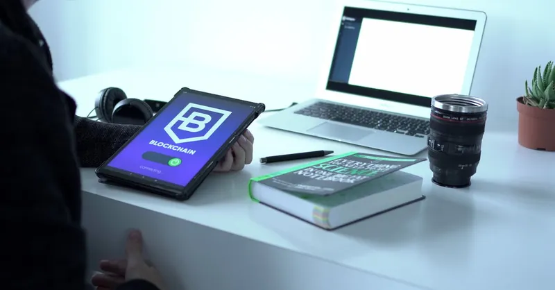A desk with tablet displaying blockchain, laptop, book, and camera lens, symbolizing tech and innovation.