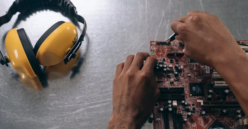 Hands repairing a motherboard on a metallic surface with yellow earmuffs beside it.