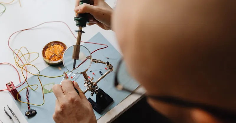 Man soldering circuit board with a magnifying glass and soldering iron in a workshop setting.